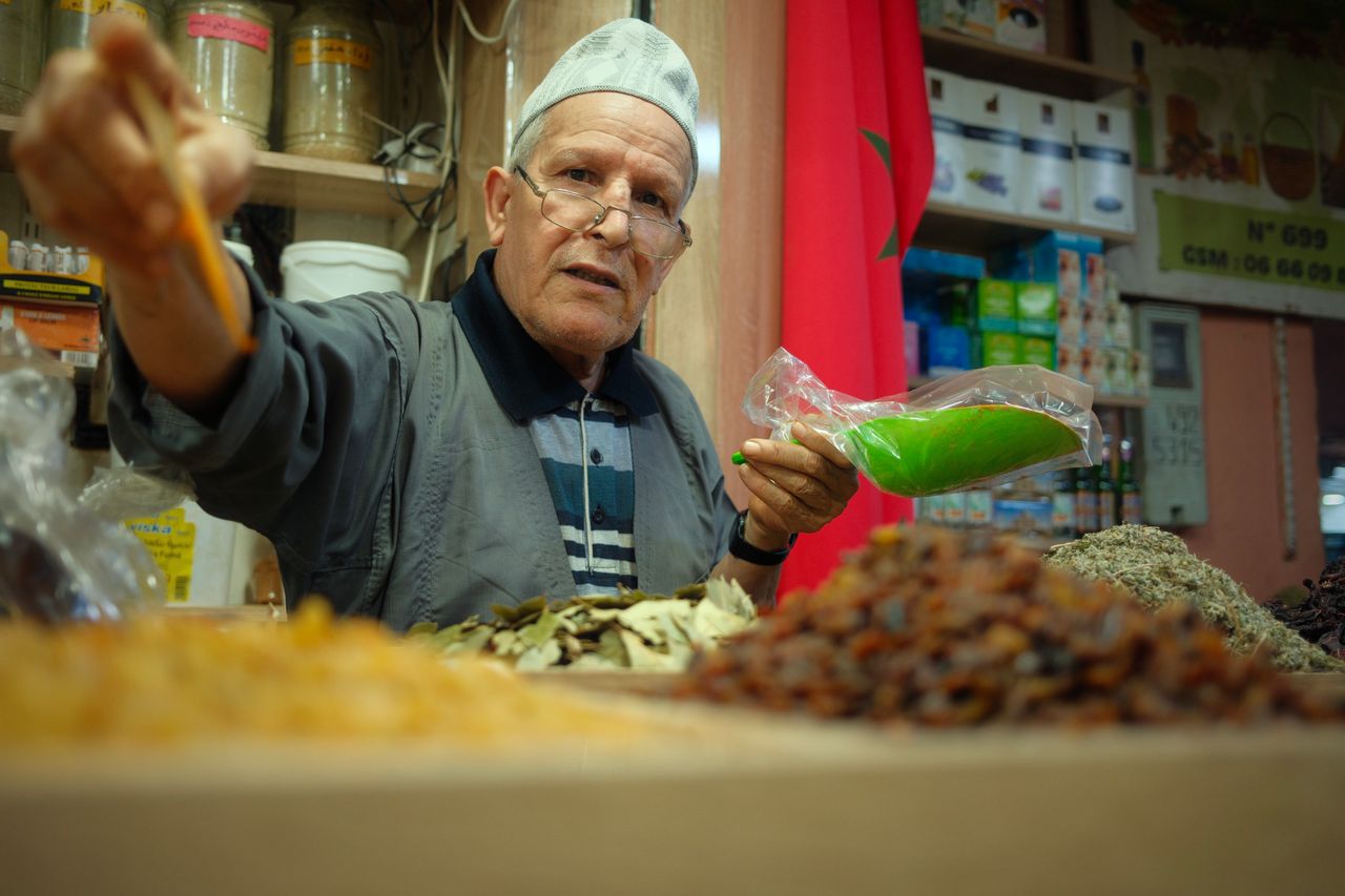 service-01 Elderly vendor at a Moroccan spice market offers vibrant spices and herbs, showcasing local culture.
