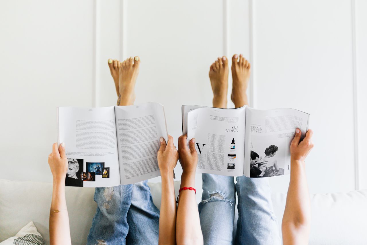 Two people reading magazines upside down in a comfortable setting with denim jeans and a white wall backdrop.